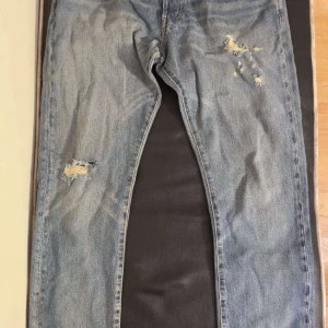 Blå jeans från Polo - Säljer ett par blå jeans från Polo med slitna detaljer och en klassisk femficksdesign. Jeansen har en straight passform och är perfekta för en avslappnad stil. De har en knappgylf och är tillverkade i denim.