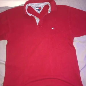 Röd pikétröja från Tommy Hilfiger - Säljer en klassisk röd pikétröja från Tommy Hilfiger med korta ärmar och en stilren krage. Tröjan har en liten broderad logotyp på bröstet och randig insida av kragen. Perfekt för en avslappnad och snygg stil.