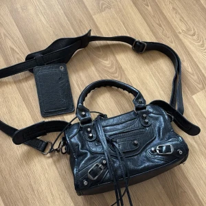 Svart axelväska med dragkedja - Intressekoll! Snygg svart axelväska i skinn med flera dragkedjor och spännen. Väskan har både handtag och en justerbar axelrem för flexibel användning. Perfekt för att bära med sig det viktigaste med stil. Säljer endast vid bra bud! Spegeln tillkommer men sitter ej fast! Finns tecken på användning 🙌