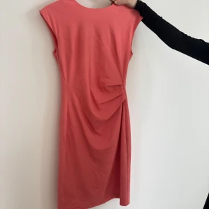 Korallfärgad klänning från Tiger of Sweden - Snygg korallfärgad klänning från Tiger of Sweden med dragkedja baktill. Klänningen har en elegant drapering på framsidan och är ärmlös, vilket ger en stilren look. Storlek 34