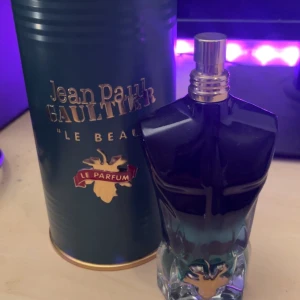 Jean Paul Gaultier Le Beau Le Parfum - Säljer en elegant parfymflaska formad som en manlig torso från Jean Paul Gaultier. Flaskan är mörkblå med en guldig detalj vid basen. Doften är intensiv och förförisk, perfekt för den som vill sticka ut.