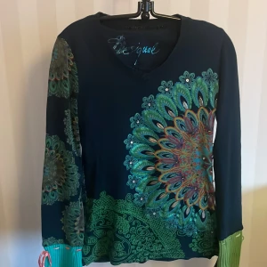 Mönstrad tröja från Desigual - Säljer en unik och färgglad tröja från Desigual med ett stort, detaljerat mönster i grönt, turkos och orange. Tröjan har långa ärmar och en v-ringad hals. Perfekt för dig som vill sticka ut med din stil!