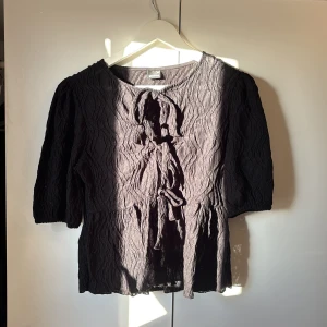 Svart topp från Gina Tricot - Snygg svart topp från Gina Tricot med knytning framtill. Toppen har trekvartsärm och ett vågigt mönster som ger en unik textur. Perfekt för en stilren look. Aldrig använd 