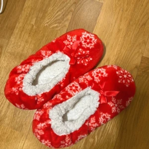 Röda tofflor med snöflingemönster - Mysiga röda tofflor med vitt snöflingemönster och en söt rosett på ovansidan. Insidan är fodrad med mjukt vitt material för extra komfort. Perfekta för kalla vinterdagar inomhus. Storlek 36, från Primark.