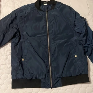 Mörkblå bomberjacka - Snygg mörkblå bomberjacka med dragkedja och ribbade muddar. Jackan har två fickor med tryckknappar och en klassisk bomberkrage. Perfekt för en stilren look.