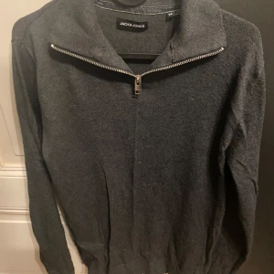 Grå tröja med dragkedja från Jack & Jones - Snygg grå tröja från Jack & Jones med dragkedja vid halsen. Perfekt för en stilren look. Tröjan har långa ärmar och är tillverkad i ett mjukt material.