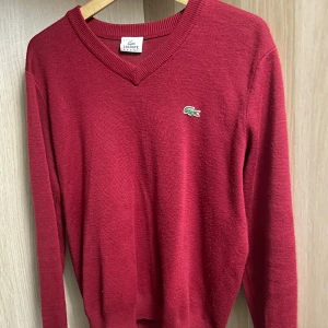 Röd tröja från Lacoste - Säljer en snygg röd tröja från Lacoste med v-ringning och det klassiska krokodilmärket på bröstet. Tröjan har långa ärmar och är perfekt för en stilren look. Passar bra till både jeans och chinos.