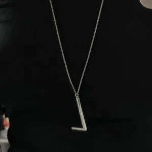 Silverfärgat halsband med L-formad hängsmycke - Elegant silverfärgat halsband med ett L-format hängsmycke dekorerat med glittrande stenar. Perfekt för att ge en stilren touch till din outfit. Kedjan är tunn och smidig, vilket gör den bekväm att bära. (Lång kedja)