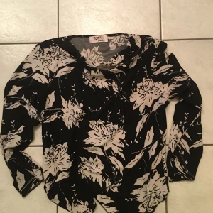 Svart blommig blus från Noa Noa - Elegant svart blus från Noa Noa med ett vackert blommönster i beige. Blusen har långa ärmar och en avslappnad passform, perfekt för en stilfull look.