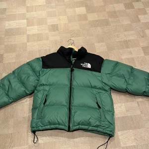 Grön och svart dunjacka från The North Face - Säljer en grön och svart dunjacka från The North Face. Jackan har en dragkedja framtill och justerbara snören i nederkant. Perfekt för kyliga dagar med sin pufferdesign och långa ärmar. Logotypen syns tydligt på bröstet och ryggen. Väldigt bra skick och har använt fåtal gånger på grund av att växt ur jackan.