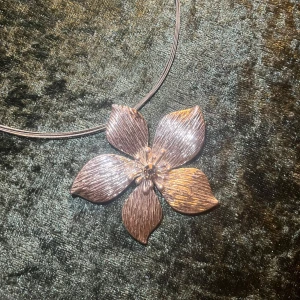 Blomformat halsband  - Elegant halsband med en stor blomma i metallisk nyans. Blomman har detaljerade kronblad och hänger på en tunn kedja. Perfekt för att ge en stilfull touch till din outfit.