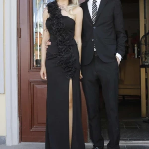 Svart långklänning från Terani Couture - Elegant svart balklänning från Terani Couture.. Klänningen har en one-shoulder design med volangdetaljer i ett rosenmönster och fjädrar samt en slits framtill som ger en sofistikerad look. Endast använd en gång så i superbra skick! Säljer endast till bra pris över 3000💕 Kom med bud! Nypris ligger runt 9-12 tusen!💕 storlek 38 men insydd så passar mig som vanligtvis har XS och S! Passar någon som är 170 och längre