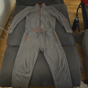 Grå tracksuit från Hugo Boss - Säljer en grå tracksuit från Hugo Boss med röda detaljer. Setet består av en tröja med dragkedja och byxor med resår i midjan. Perfekt för en avslappnad dag.