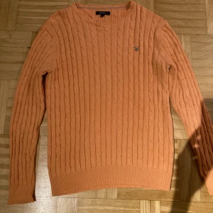 Orange kabelstickad tröja från GANT - Säljer en snygg orange stickad tröja från GANT med kabelstickat mönster. Tröjan har långa ärmar och ribbade muddar vid ärmslut och nederkant. Perfekt för kyligare dagar!