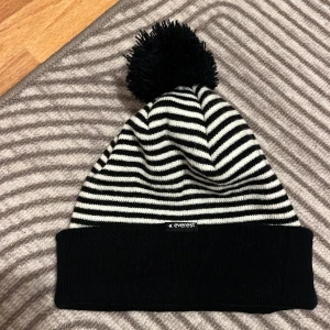 Randig mössa med tofs från Everest - Snygg svart och vit randig mössa från Everest med en fluffig tofs på toppen. Perfekt för kyliga dagar med sin stickade design. Märkt med Stadium. One size storlek