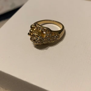 Guldfärgad ring med leoparddesign - Snygg guldfärgad ring med en leoparddesign prydd med glittrande stenar. Perfekt för att ge en djärv touch till din stil. Ringen har en unik och iögonfallande design som verkligen sticker ut. Köpt den begagnad så vet inte märke osv. Vikt 7 gram, storlek 17,7. 