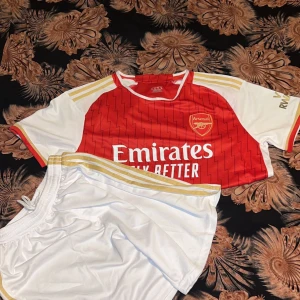 Röd Arsenal fotbollströja - Säljer en snygg röd Arsenal fotbollströja från Adidas med vita och guldiga detaljer. Tröjan har korta ärmar och ett subtilt randigt mönster. Perfekt för alla Arsenal-fans! 🔴⚪️