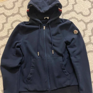 Mörkblå hoodie från Moncler - Säljer en snygg mörkblå hoodie från Moncler med dragkedja och huva. Den har ett stilrent märke på ärmen och röd-vita detaljer på huvan. Perfekt för en casual look.
