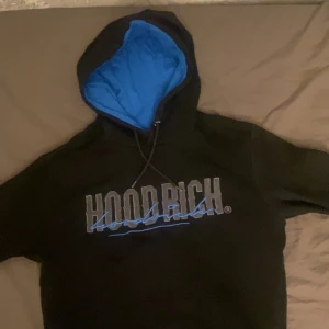 Svart hoodie från Hoodrich - Säljer en svart hoodie från Hoodrich med blått foder i huvan och tryck på bröstet. Perfekt för streetwear-stilen. Huvan har dragsko för justering.