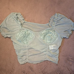 Blågrön topp med snäckdetaljer från Romwe - Säljer en blågrön topp från Romwe med unika snäckdetaljer på bröstet. Toppen har korta puffärmar och en smickrande passform. Perfekt för en somrig look! Endast testad. 💙