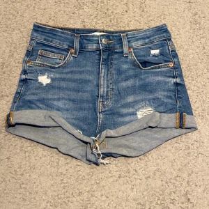 Blå jeansshorts från H&M - Snygga blå jeansshorts från H&M med slitna detaljer och uppvikta ben. Perfekta för en avslappnad stil. De har klassisk femficksdesign och knappgylf.
