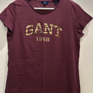 Vinröd t-shirt från GANT - Säljer en snygg vinröd t-shirt från GANT med guldtryck av märkesnamnet och årtalet 1949 på framsidan. T-shirten har en rund halsringning och korta ärmar. Perfekt för en avslappnad stil.