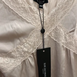 Ilse Jacobsen nattlinne - Elegant champagnefärgad klänning/nattlinne från Ilse Jacobsen med vackra spetsdetaljer vid halsringningen och fållen. Klänningen är axelbandslös och har en mjuk, glansig yta som ger en lyxig känsla. Perfekt för speciella tillfällen.