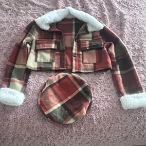 Rutig croppad jacka med fuskpäls - Säljer en supercool rutig croppad jacka med fuskpälsdetaljer på krage och ärmslut. Jackan har en härlig mix av röd, beige och brun färg och två fickor framtill. Perfekt för att ge din outfit en unik touch!