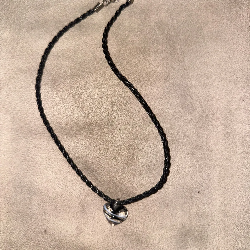 Snyggt svart flätat halsband med ett hjärthänge i glas. Hänget har ett unikt mönster i svart och vitt. Halsbandet har en praktisk karbinhake för enkel på- och avtagning.. Asusteet.