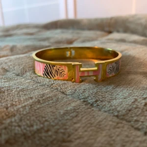 Guldarmband med zebramönster - Elegant guldarmband med ett unikt zebramönster i rosa och svart. Armbandet har en distinkt H-formad detalj i mitten som ger det en exklusiv touch. Perfekt för att lägga till en färgklick och stil till din outfit.