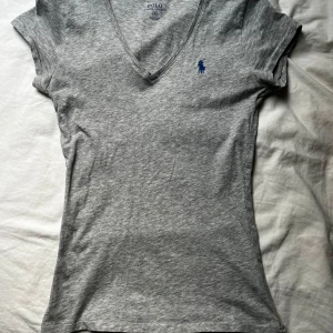 Grå v-ringad t-shirt från Ralph Lauren - Säljer en stilren grå t-shirt från Ralph Lauren med v-ringning och korta ärmar. Den har en liten blå logga på bröstet. Perfekt för en avslappnad look.