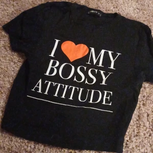 Svart t-shirt med tryck - Cool svart t-shirt från new yorker  med texten 'I ❤️ MY BOSSY ATTITUDE' i vitt och orange. Perfekt för att uttrycka din personlighet med stil. Kortärmad och bekväm passform.