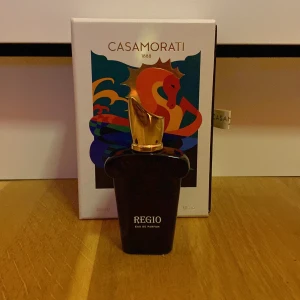 Regio Eau de Parfum från Casamorati - Elegant parfymflaska från Casamorati med en unik design. Flaskan är mörk med en gyllene kork formad som en stiliserad hand. Förpackningen har en färgglad illustration med en drake. Perfekt för den som söker en lyxig doftupplevelse.