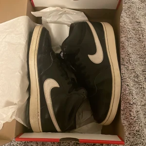 Svarta Nike sneakers med vit sula - Snygga svarta Nike sneakers med vit sula och klassisk Nike-logga i vitt på sidan. Skorna har snörning och är tillverkade i skinn, vilket ger en stilren look. Perfekta för en avslappnad stil. Är slitna på utseendet men kommer hålla bra ändå i längden.