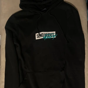 Svart hoodie från FSBN - Säljer en svart hoodie från FSBN med texten 'SEND NUDES' överstruket och ersatt med 'MONEY' i turkos. Hoodien har en klassisk design med dragsko i huvan och en stor ficka framtill. Perfekt för en avslappnad stil.