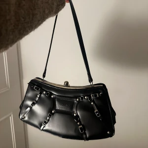 Svart axelväska  - Snygg svart axelväska med unika spänndetaljer och en elegant metallknäppning. Perfekt för att ge din outfit en stilfull touch. 