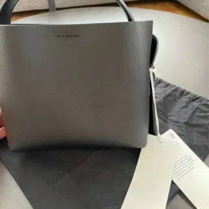 Svart handväska från ARKET - Snygg svart handväska från ARKET  i elegant design. Tillverkad i skinn med stilrena linjer och en justerbar axelrem. Perfekt för att ge en touch av lyx till din outfit. Väskan har en minimalistisk look med subtila detaljer.   Magnet förslutning och innerficka. Ej använd.  Mått : 14 cm x 17 cm x 6 cm.