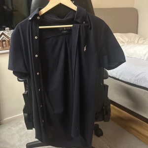 Ralph lauren skjorta - Snygg mörkblå kortärmad skjorta från Ralph Lauren och den är lite lång i modell. Jag köpte den i jhonells för cirka 1800kr men har ingen typ av ektihets bevis. 