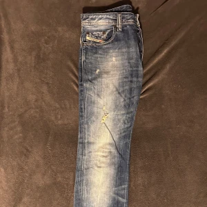 Blå diesel Jean med slitningar  - Snygga blå jeans med slitningar och en klassisk femficksdesign. De har en knappgylf och är i en straight passform. Perfekta för en avslappnad stil. Storlek W29 L34