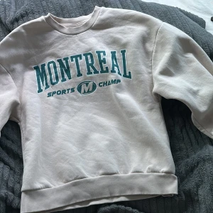 Vit sweatshirt med tryck - Säljer en vit sweatshirt med texten 'Montreal Sports Champ' i grönt tryck. Tröjan har en klassisk rund hals och långa ärmar. Perfekt för en avslappnad stil.