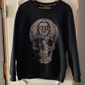 Svart tröja med dödskalle från Philipp Plein - Säljer en svart tröja från Philipp Plein med en stor, glittrig dödskalle på framsidan. (Replika) Tröjan har långa ärmar och en rund halsringning. Perfekt för dig som gillar en edgy stil. 🖤 storleksmässigt passar som en Large!