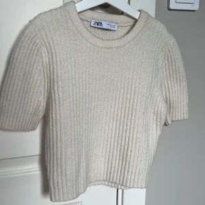 Beige ribbad tröja från Zara - Säljer en stilren stickad beige ribbad tröja från Zara. Tröjan har korta ärmar och en rund halsringning. 