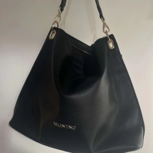 Svart handväska från Valentino - Snygg svart axelväska från Valentino i skinn. Väskan har en stilren design med logotypen diskret placerad på framsidan. Perfekt för skolan eller annat vardagligt💕 Knappt använd, köptes för 1500kr, säljer då den inte kommer till användning ❣️