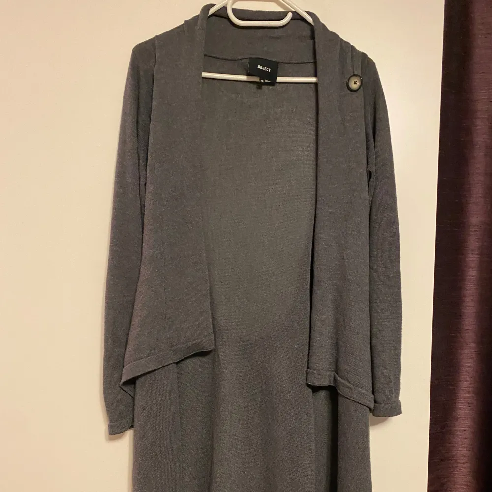 Grå lång kofta från Object i strl xs (passar xs-m) med knytning i ryggen. Koftan har en stilren design med en knappdetalj vid axeln (på tredje bilden är den knäppt) och en mjuk, bekväm passform. . Neuletakit & Villapaidat.