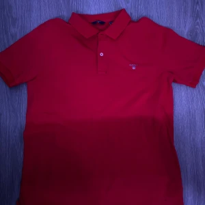 Röd pikétröja från Gant - Säljer en klassisk röd pikétröja från Gant. Tröjan har korta ärmar och en traditionell krage med två knappar. Perfekt för en stilren och avslappnad look.