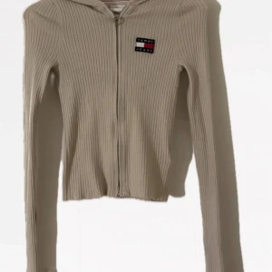 Beige ribbad hoodie från Tommy Jeans - Säljer en beige ribbad hoodie från Tommy Jeans med dragkedja och logga på bröstet. Tröjan är croppad och har långa ärmar. Perfekt för en avslappnad stil. En liten fläck.