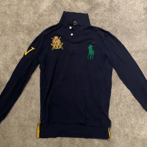 Mörkblå pikétröja från Polo Ralph Lauren - Snygg mörkblå pikétröja från Polo Ralph Lauren med grönt broderat logotyp på bröstet och ett detaljerat emblem på andra sidan. Tröjan har långa ärmar och en klassisk krage med knappar. Perfekt för en stilren look.