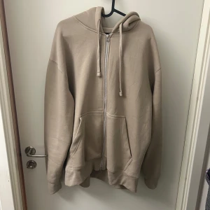 Beige hoodie med dragkedja - Säljer en stilren beige hoodie med dragkedja och justerbar huva. Perfekt för en avslappnad look. Den har långa ärmar och praktiska fickor framtill.