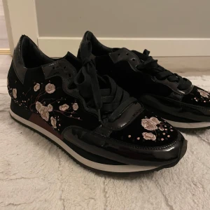 Svarta sneakers med blommigt mönster - Snygga svarta sneakers från Philippe Model med blommigt broderi i rosa och beige. Skorna har en glansig finish och snörning framtill. Perfekta för att ge en stilfull touch till din outfit.