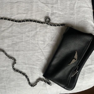 Svart axelväska med kedja - Snygg svart axelväska i skinn med en cool kedjerem. Väskan har en dragkedja och en dekorativ detalj på framsidan. Perfekt för att ge din outfit en edgy touch.